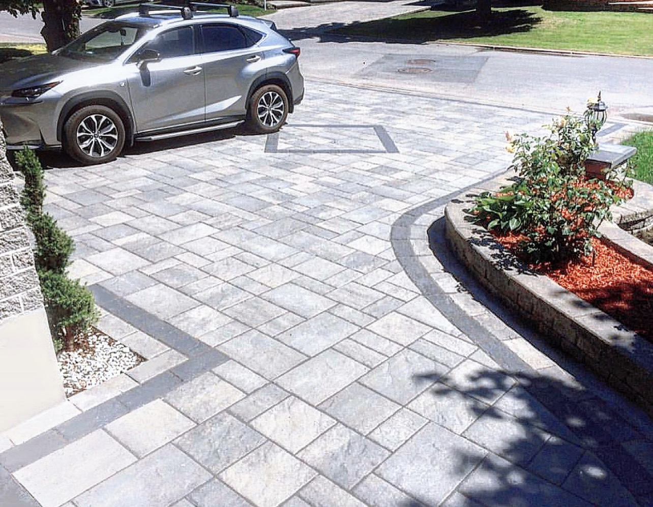 Grey interlocking driveway with a dark border, stone steps and landscaped beds beside a stone home
