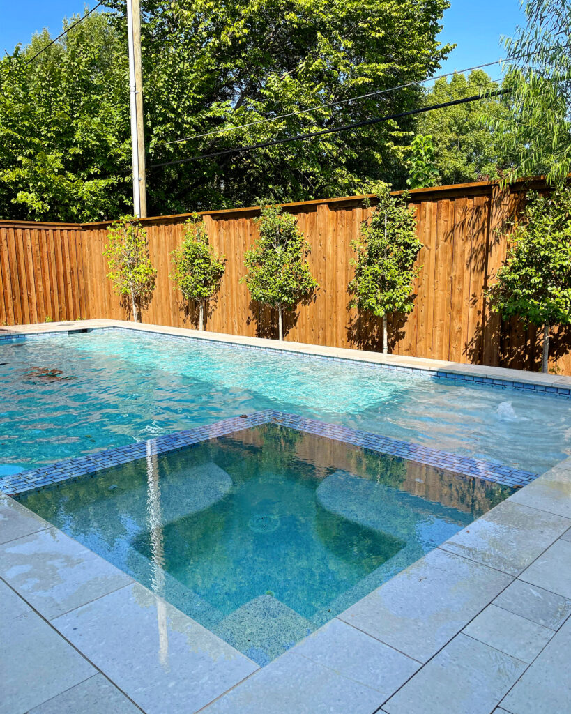Rectangular swimming pool with integrated spa, blue tile trim, light stone coping, and natural cedar privacy fence
