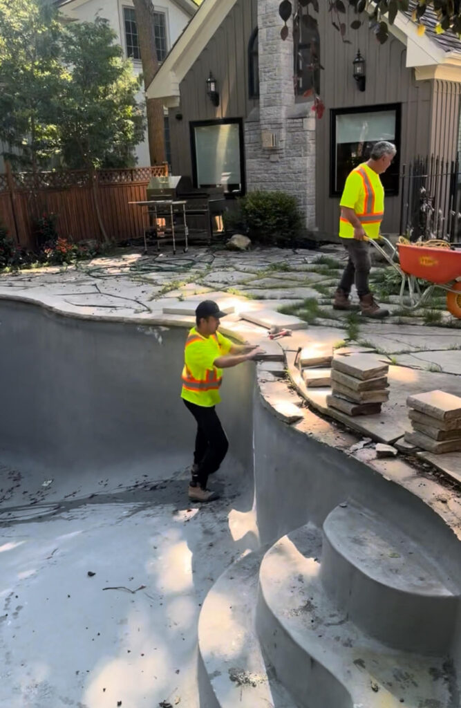 Workers in safety vests installing curved concrete pool steps and coping beside a modern backyard with stone patio.