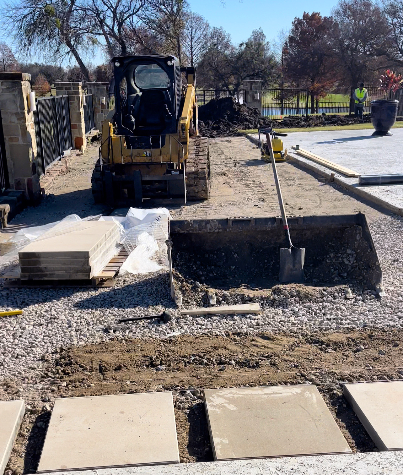 Construction site showing compact track loader, excavated foundation area with gravel base, stacked pavers, and work in progress.