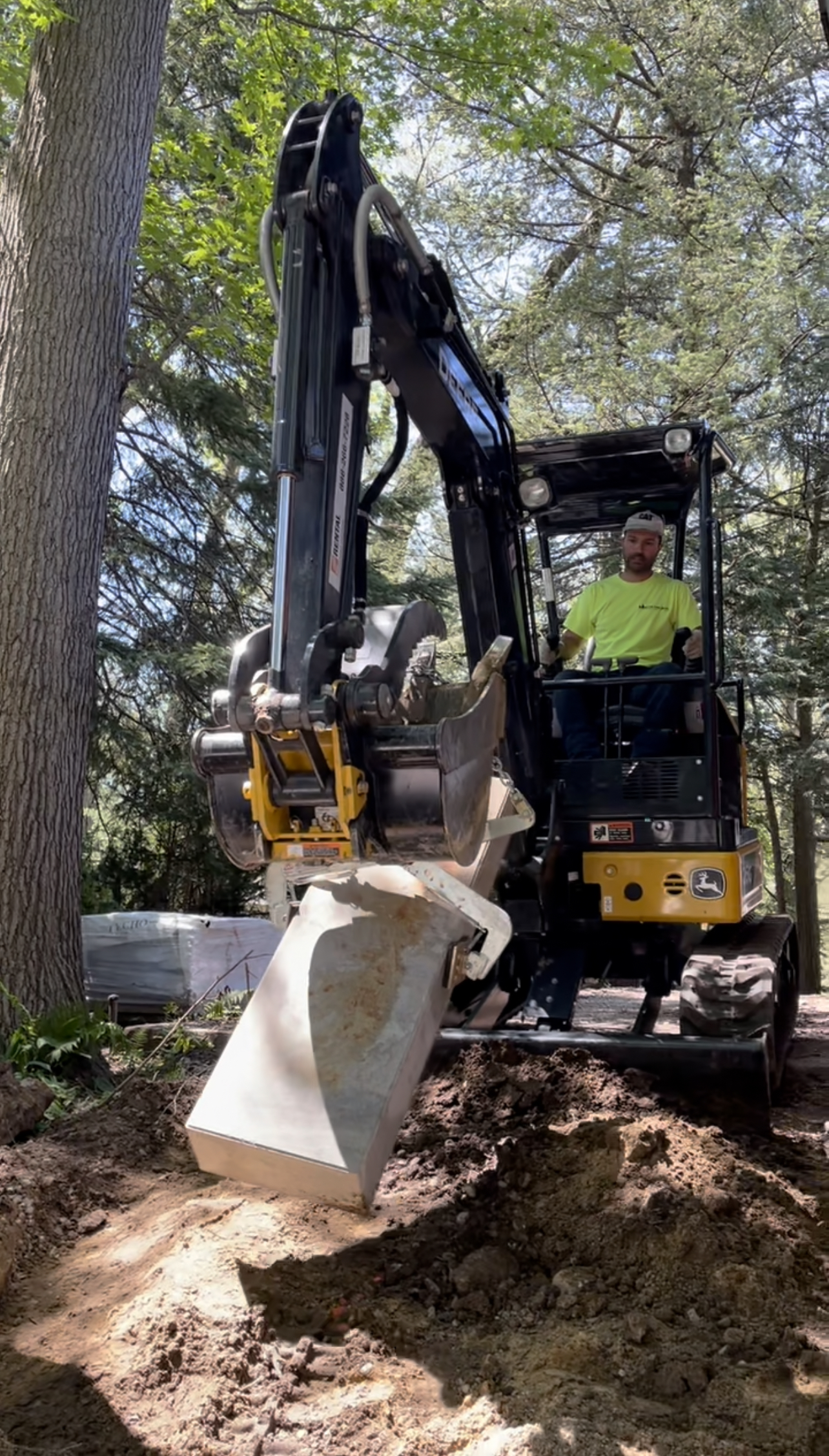 Compact excavator with yellow bucket lifting dirt from excavation site surrounded by trees during landscaping project