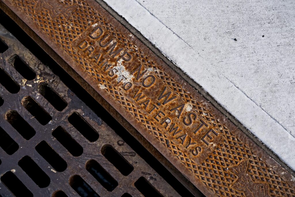 pexels-photo-6573773-6573773 Close-up of a rusty drainage grate with environmental warning text on concrete sidewalk