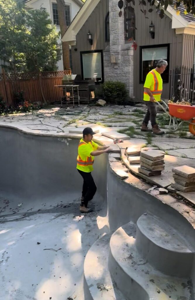 Two workers in safety vests installing fiberglass pool shell with curved steps and stone coping in progress