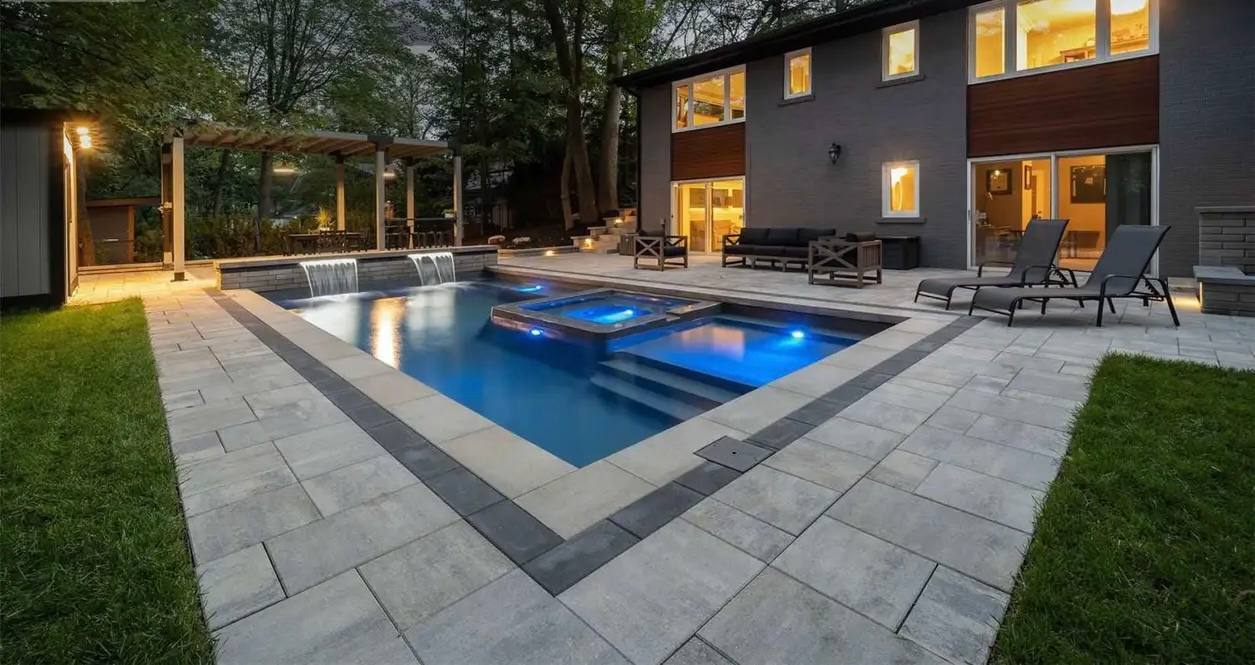 Modern rectangular pool with integrated hot tub, blue LED lighting, gray stone pavers with dark border accent, and contemporary house backdrop at dusk