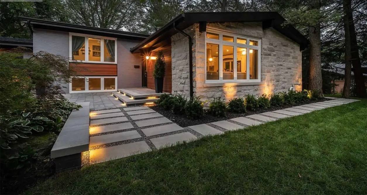 Modern home exterior with large format concrete pavers set in lawn, uplighting on stone facade, and illuminated entry steps at dusk.