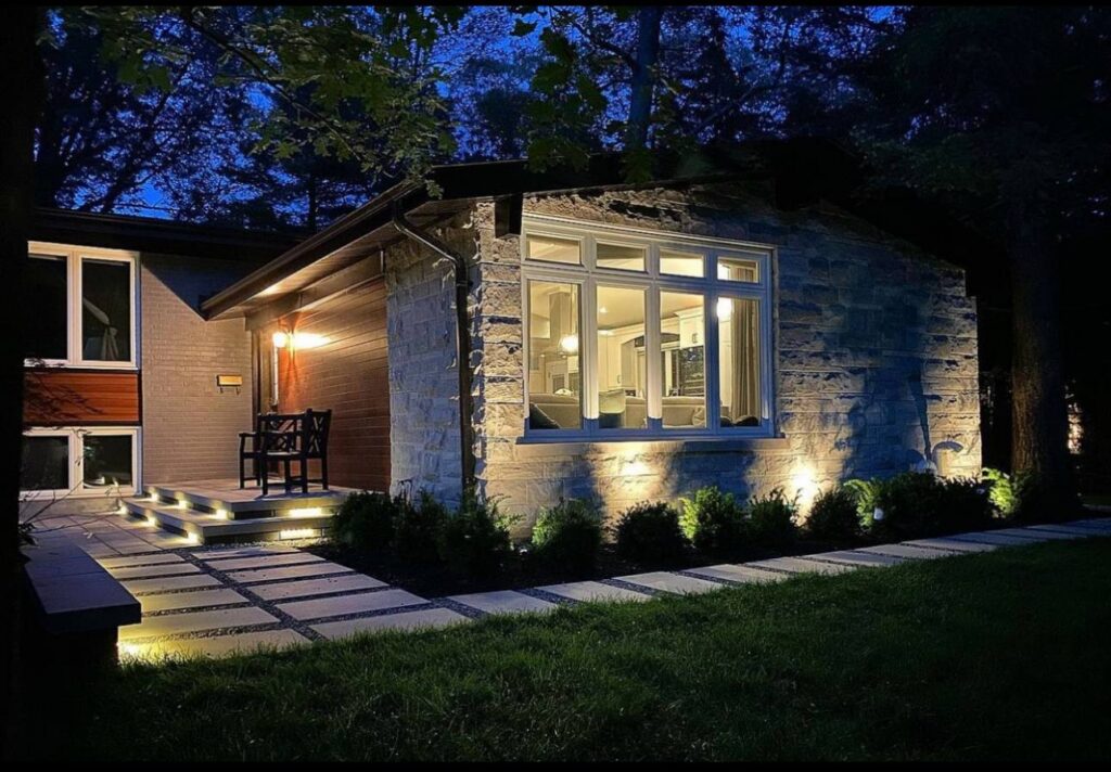 Modern home exterior at dusk with stacked stone facade, large windows, uplighting, and stone paver pathway with integrated LED lights.