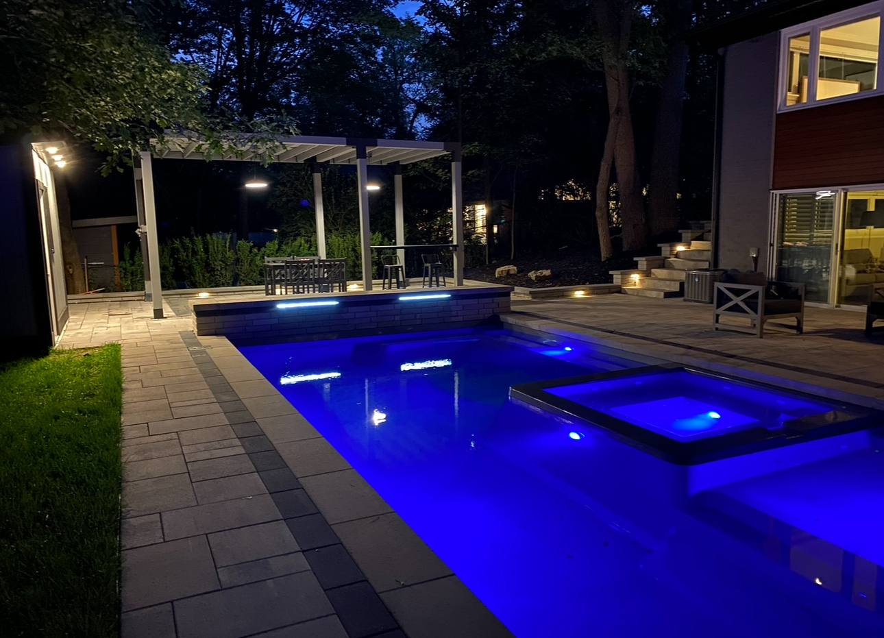 Luxury rectangular pool with blue LED lighting, raised spa, paver deck, white pergola, and outdoor kitchen at dusk