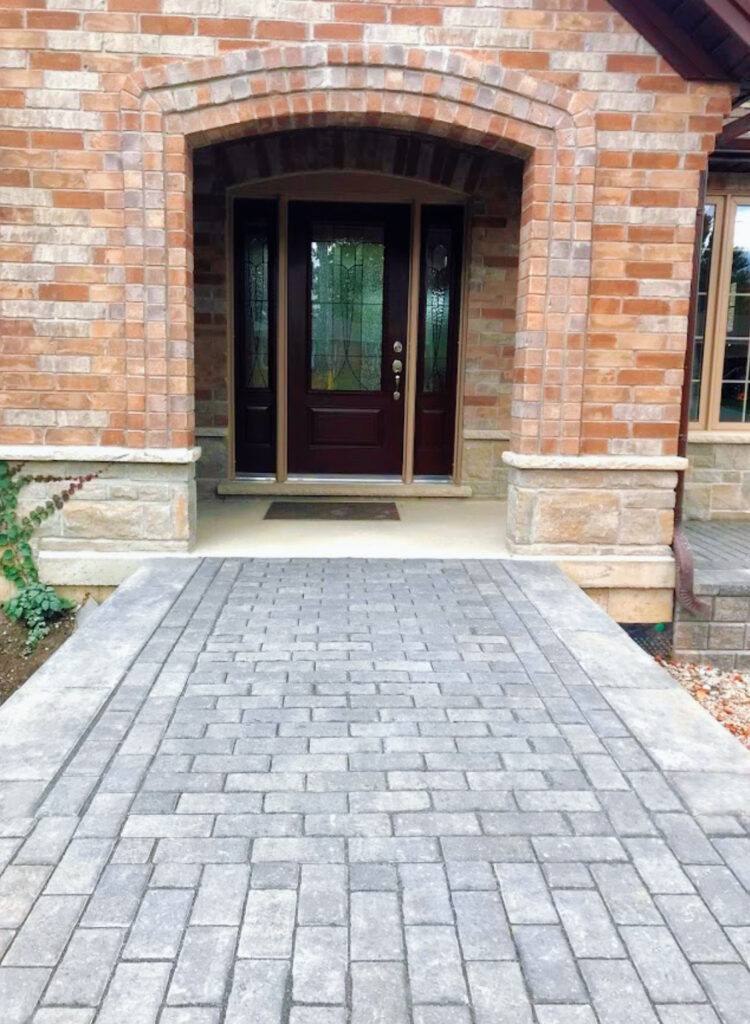 Interlocking paver walkway in running bond pattern leading to brick home entrance with arched black door and stone pillars