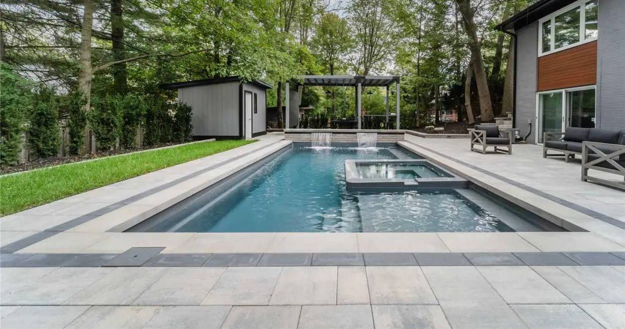 Modern rectangular pool with raised spa, bordered by light and dark grey pavers, surrounded by lush lawn and mature trees.