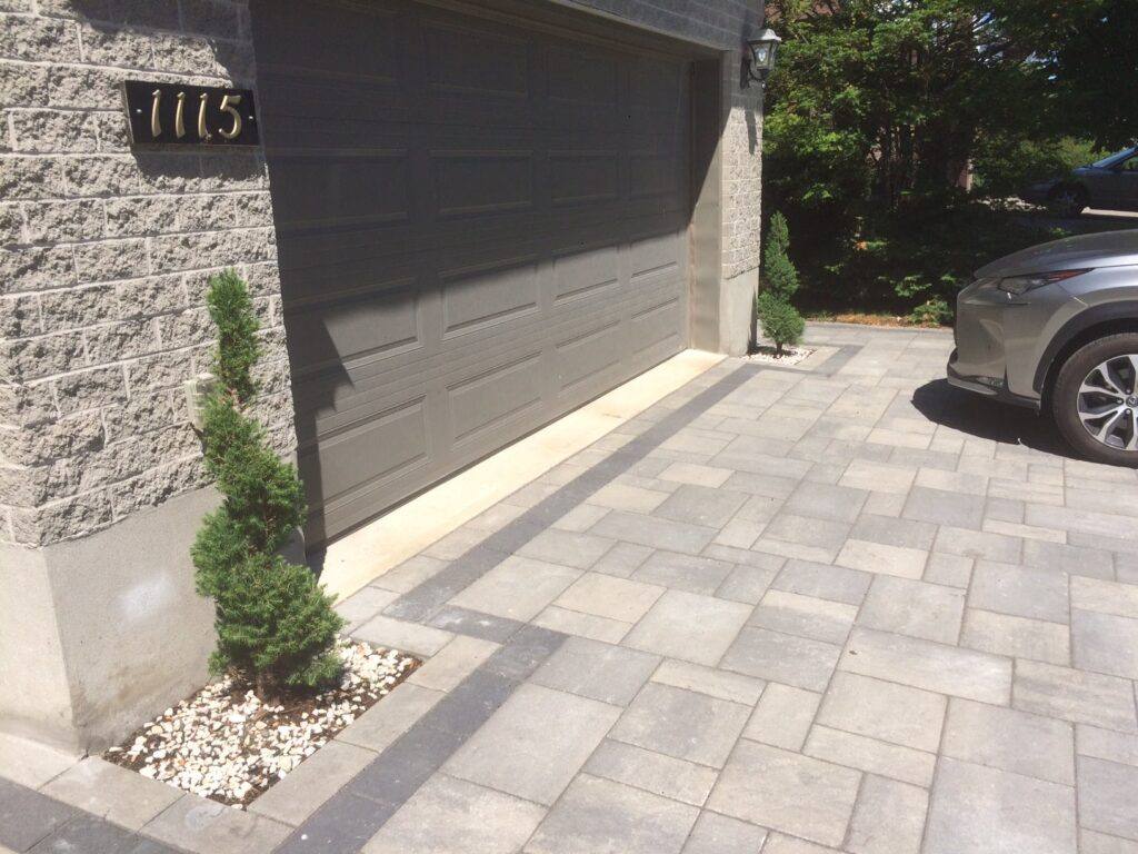 Interlocking paver driveway in rectangular pattern with darker border course leading to garage and evergreen shrub planting bed