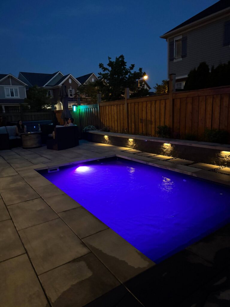 Rectangular swimming pool with purple LED lighting, paver deck, wood privacy fence, hot tub and step lighting at dusk