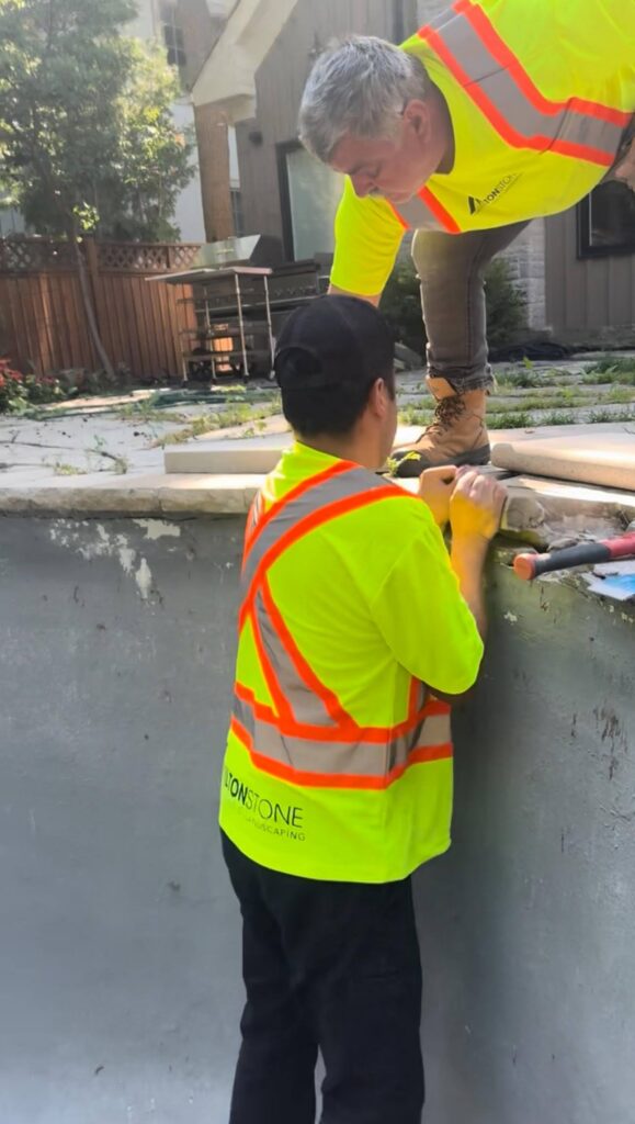 Two workers in safety vests installing concrete pool coping on a construction site with overgrown pavers visible behind them