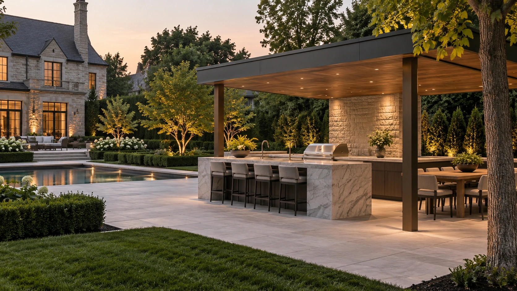 Covered outdoor kitchen with a stone island beside a pool and mature estate garden